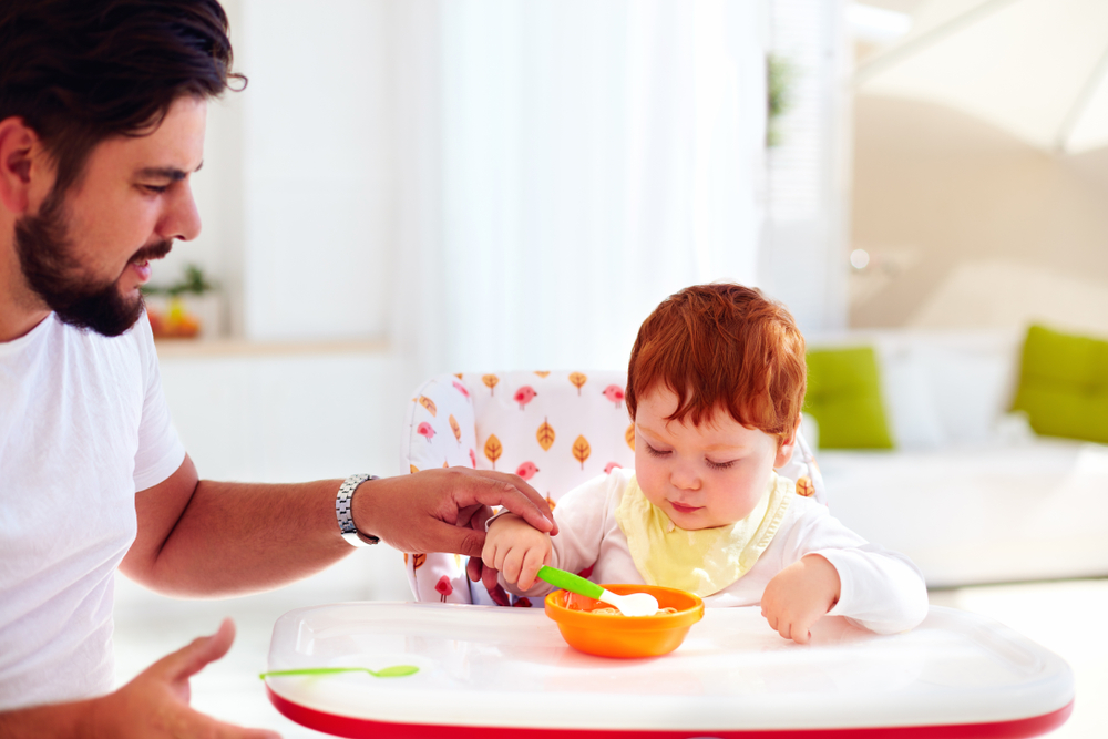 Papà che insegna al suo bambino sul seggiolone a mangiare da solo aiutandolo a tenere il cucchiaio