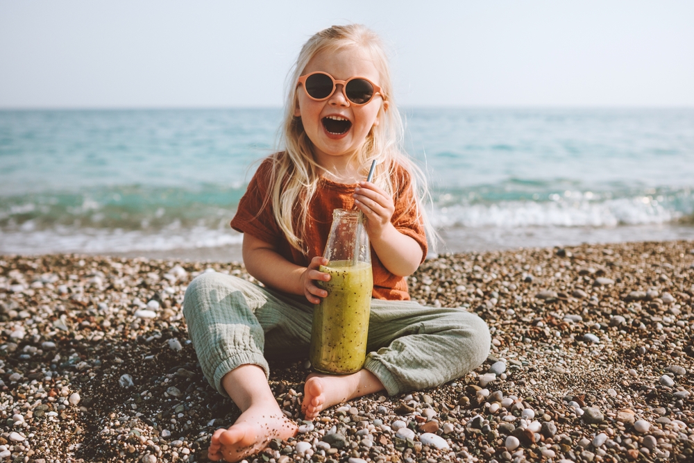 Bambina sorridente in riva al mare che mangia uno smoothie