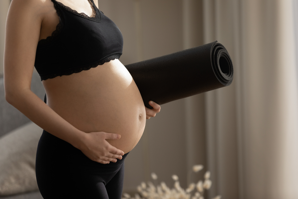 Mamma in top e pantaloncini neri che si accarezza il pancione con la mano destra e tiene il tappetino arrotolato sotto il braccio sinistro