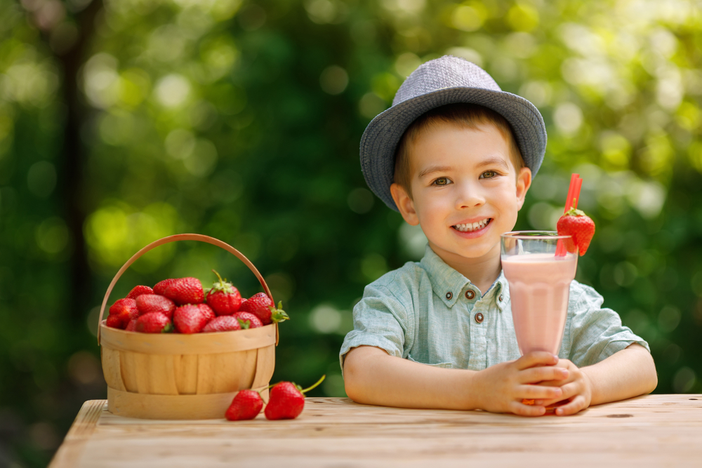 Bambino sorridente seduto al tavolo con in mano un bicchiere con un frullato alle fragole e alla sua sinistra un cestino di fragole