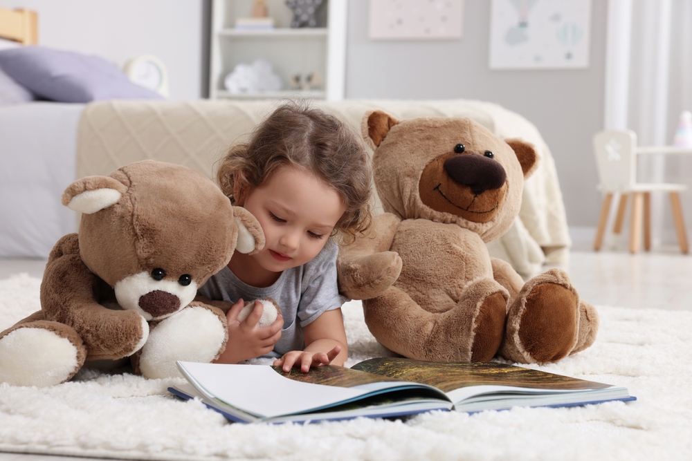 Bambina sdraiata sul tappeto con due peluche a forma di orso che legge un libro