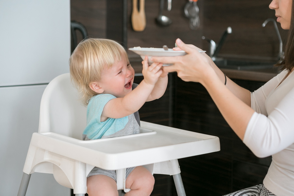 Bambino sul seggiolone che piange e spinge il piatto della mamma rifiutando il cibo