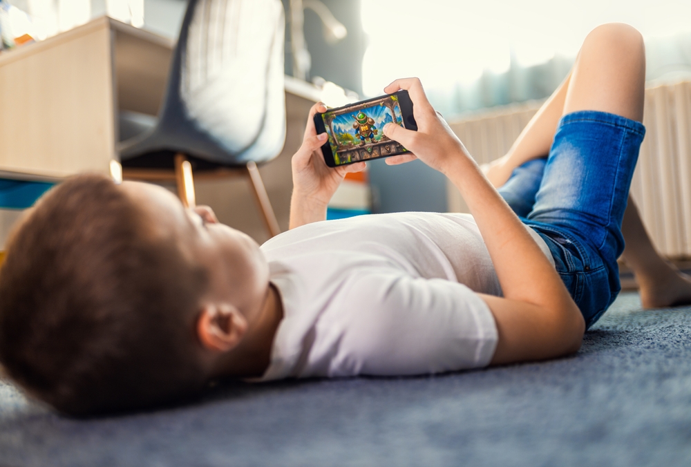 Bambino sdraiato sul pavimento della sua stanza mentre gioca a un videogioco sullo smartphone