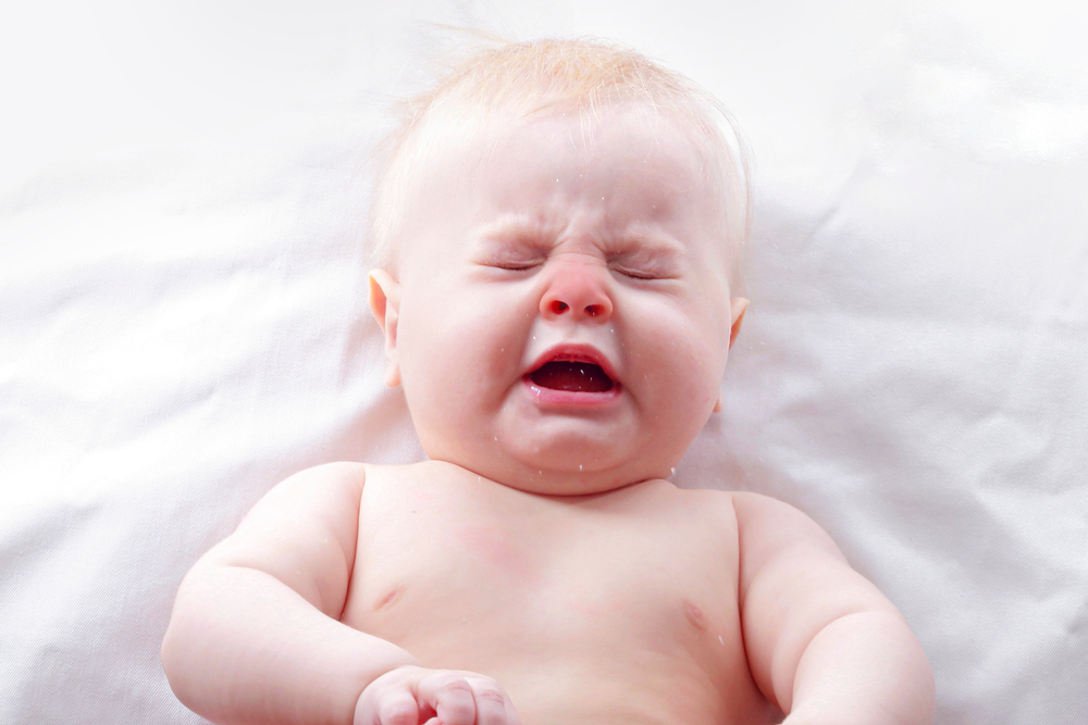 Neonato sdraiato su un lenzuolo bianco mentre starnutisce, con il viso contratto e gli occhi chiusi