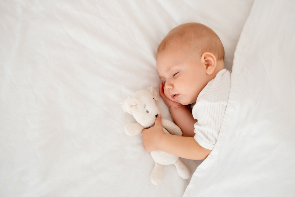 Neonato addormentato che abbraccia un orsetto di peluche sotto una coperta bianca