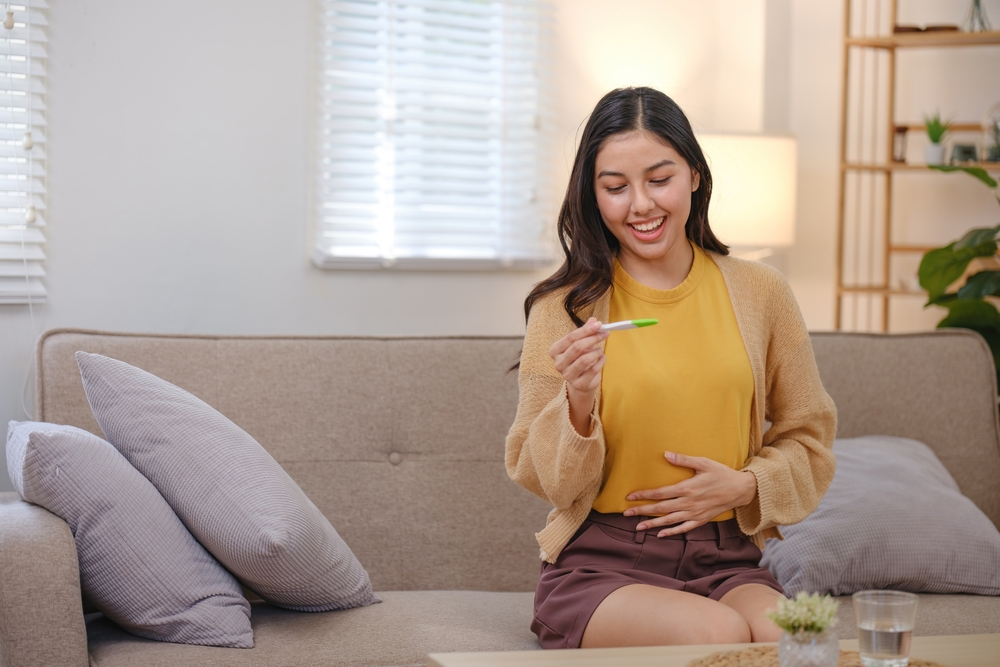 Donna seduta sul divano con una mano sul ventre e l’altra che tiene un test di gravidanza, mentre guarda il risultato