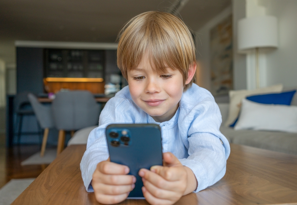 Bambino sorridente sdraiato sul tavolo della cucina mentre interagisce con lo smartphone